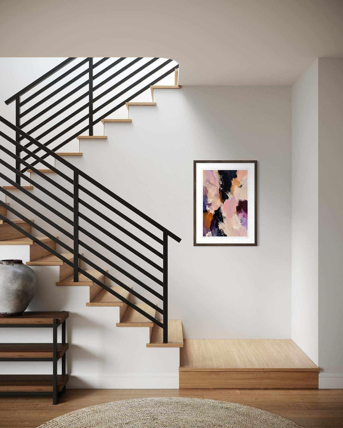 A framed abstract art print with pink, purple, and black brushstrokes hangs on a white wall in a modern staircase hallway with light wood steps and black metal railings.
