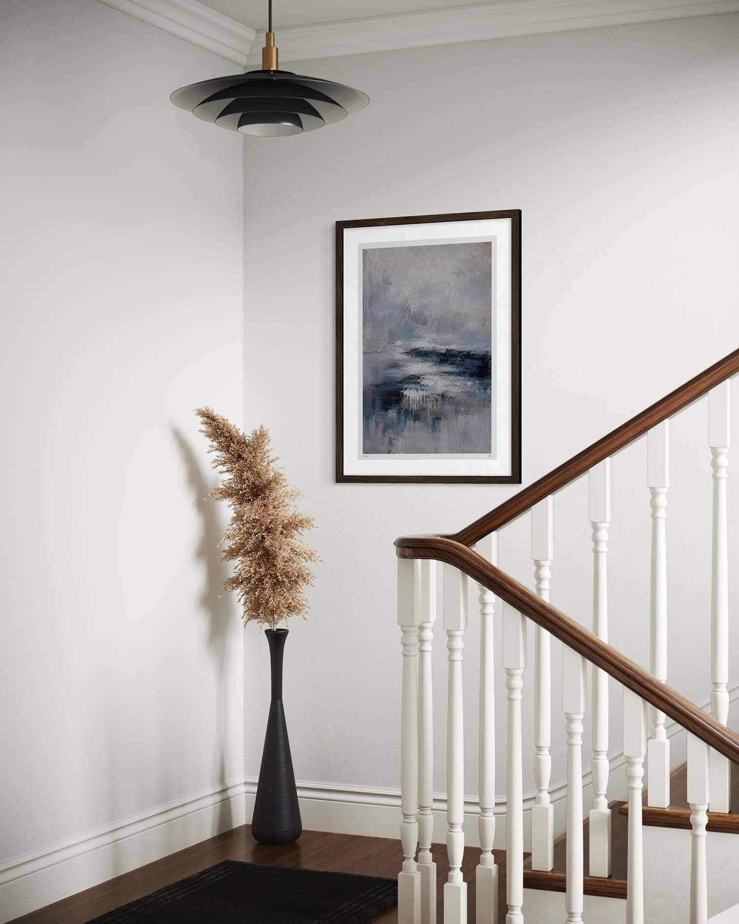 A framed abstract landscape art print with blue, gray, and white tones hangs on a white wall in a staircase landing. The scene includes a wooden handrail with white balusters, a black vase with dried pampas grass, and a black pendant light.