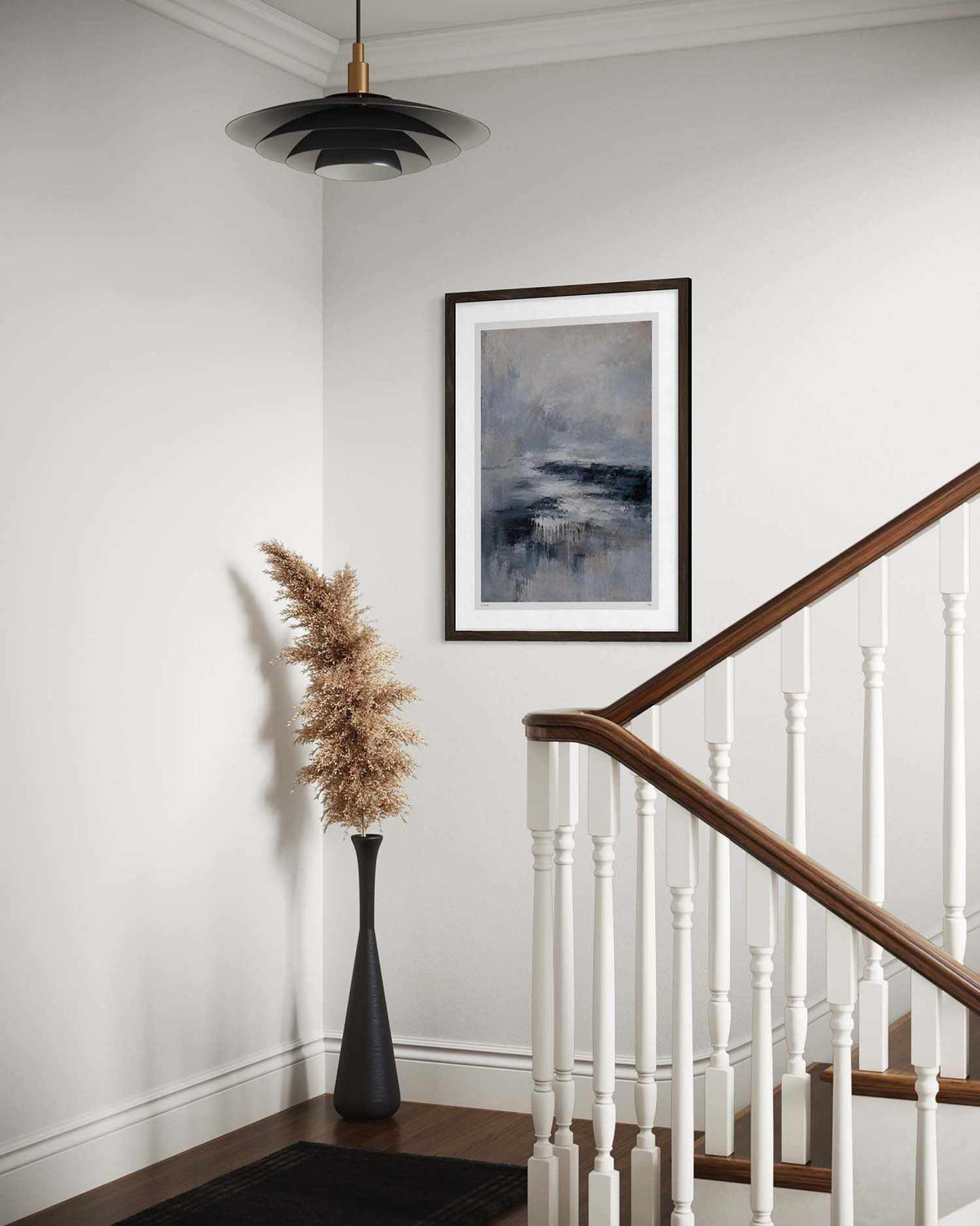A framed abstract landscape art print with blue, gray, and white tones hangs on a white wall in a staircase landing. The scene includes a wooden handrail with white balusters, a black vase with dried pampas grass, and a black pendant light.