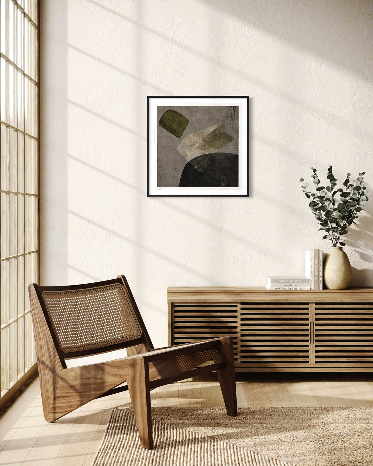 A framed abstract art print hangs on a textured cream wall in a modern, sunlit room. The print features a minimalist composition with muted gray, olive green, and black shapes.