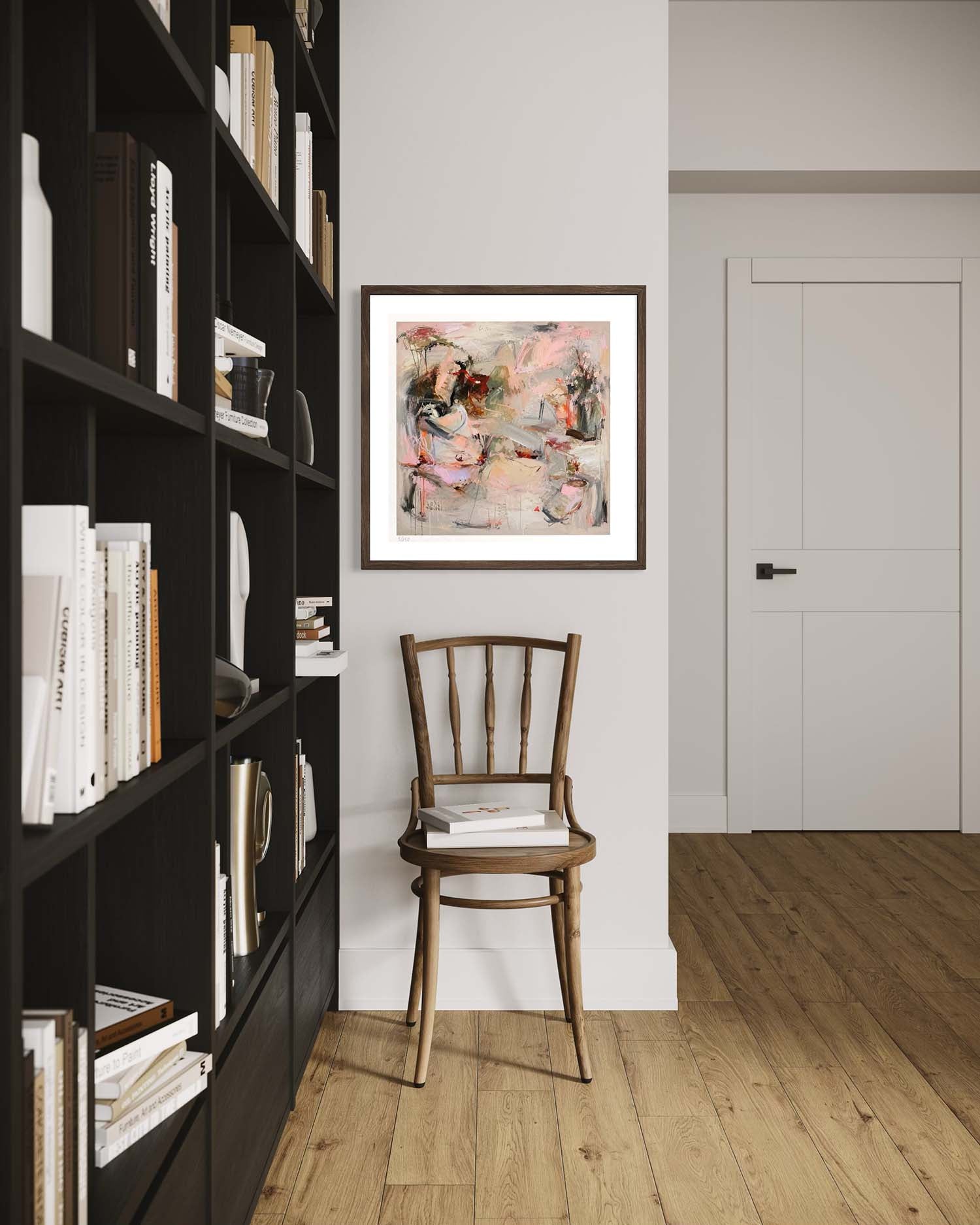 A framed abstract painting with pink, beige, and gray tones hangs on a white wall in a modern room with a wooden chair, a tall black bookshelf, and light wood flooring.