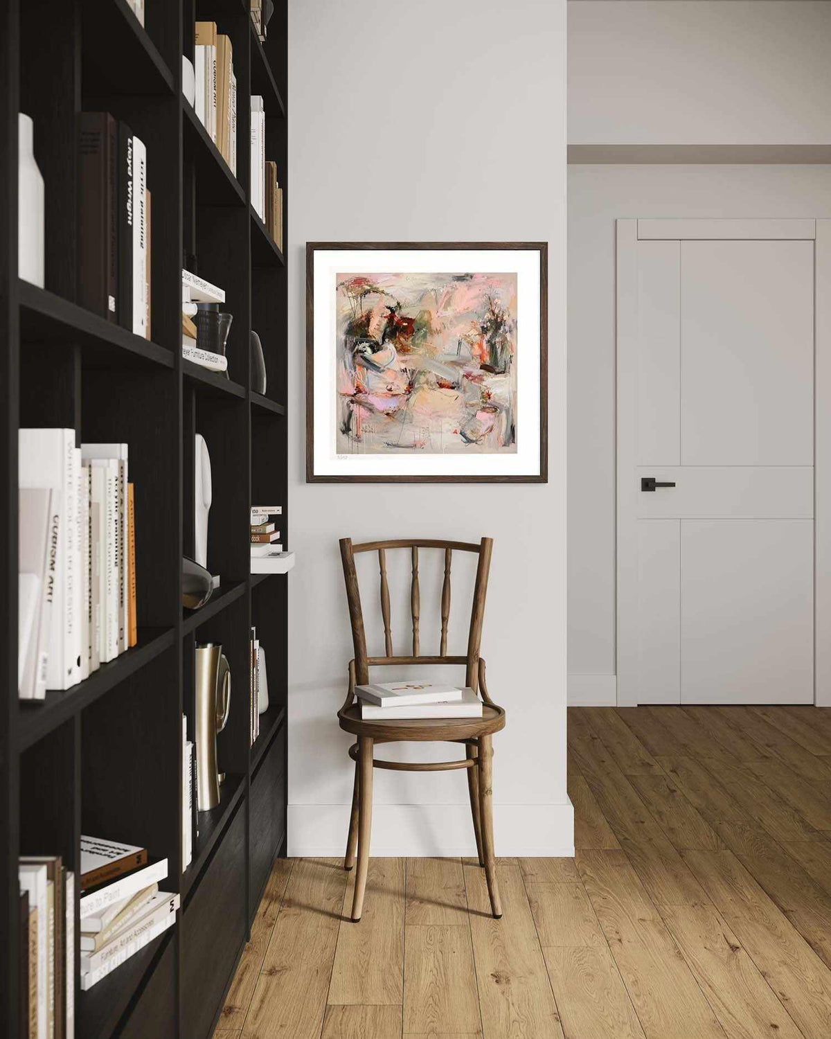 A framed abstract painting with pink, beige, and gray tones hangs on a white wall in a modern room with a wooden chair, a tall black bookshelf, and light wood flooring.