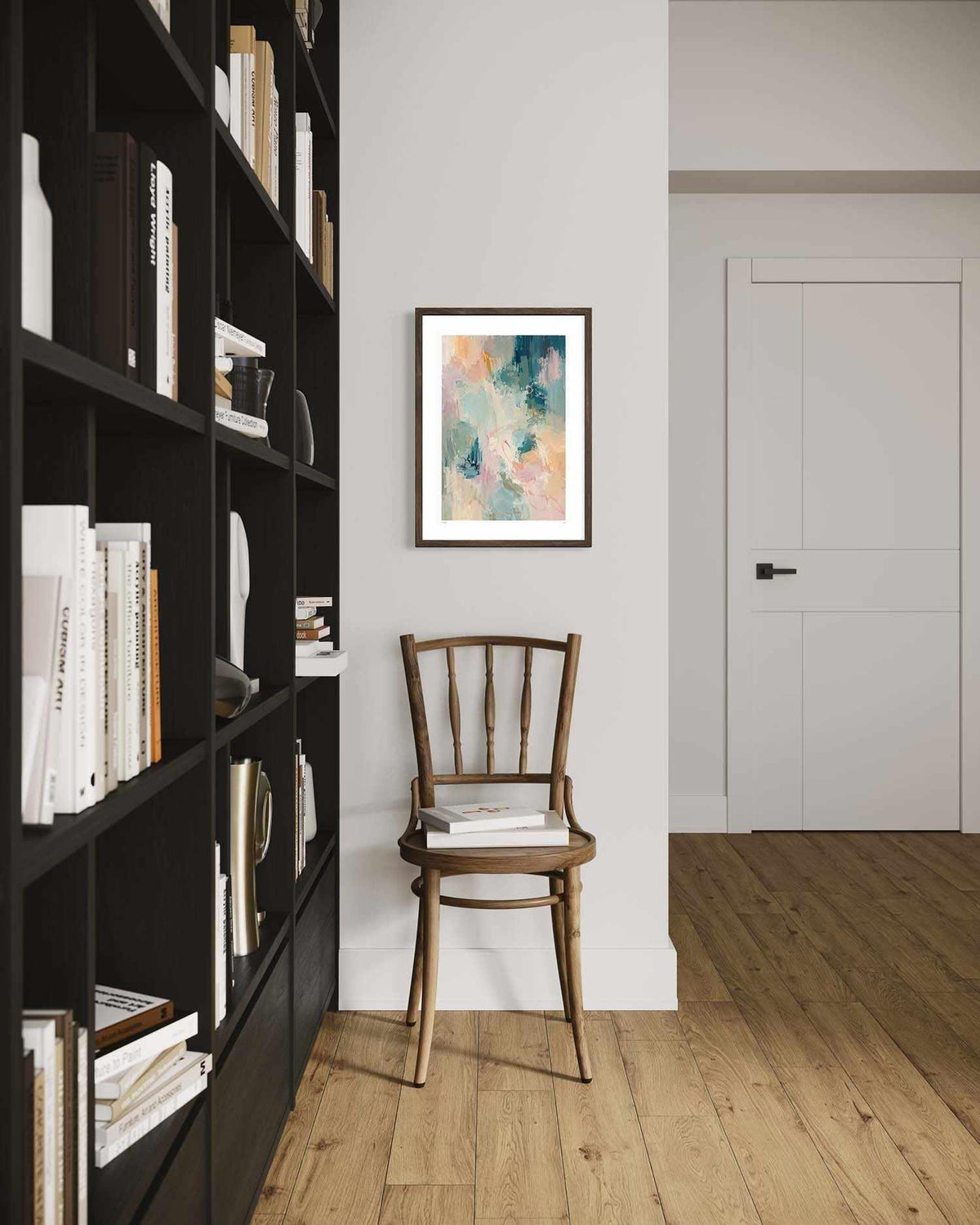 A framed abstract art print with teal, pink, and cream tones hangs on a white wall in a modern room setting with a wooden chair and a tall black bookshelf.