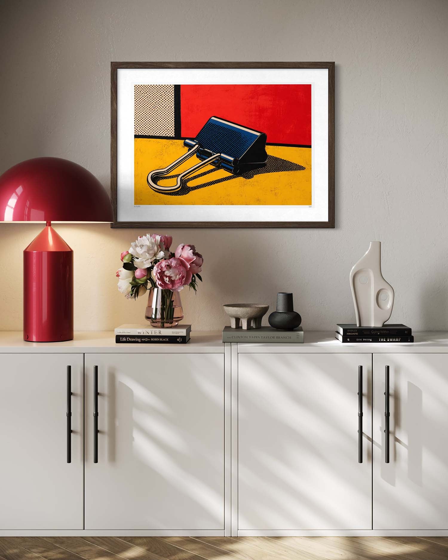 A framed abstract art print featuring a binder clip on a yellow and red background, displayed on a light gray wall above a white sideboard with a red lamp, flowers, and decorative objects.