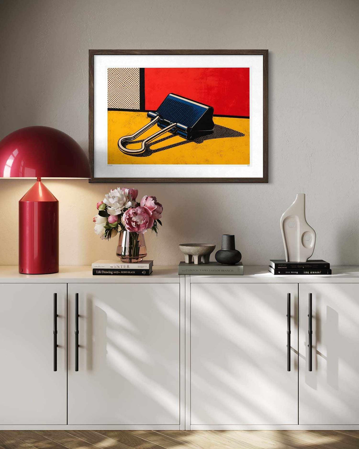A framed abstract art print featuring a binder clip on a yellow and red background, displayed on a light gray wall above a white sideboard with a red lamp, flowers, and decorative objects.