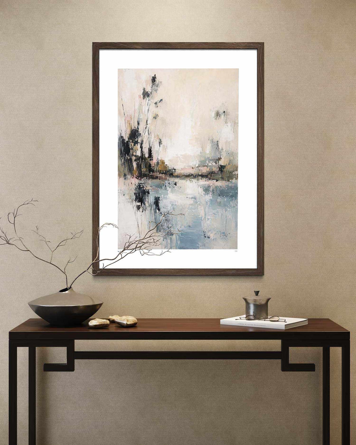 A framed abstract landscape art print with blue, gray, and beige tones, displayed on a textured beige wall above a dark wood console table with decorative items.
