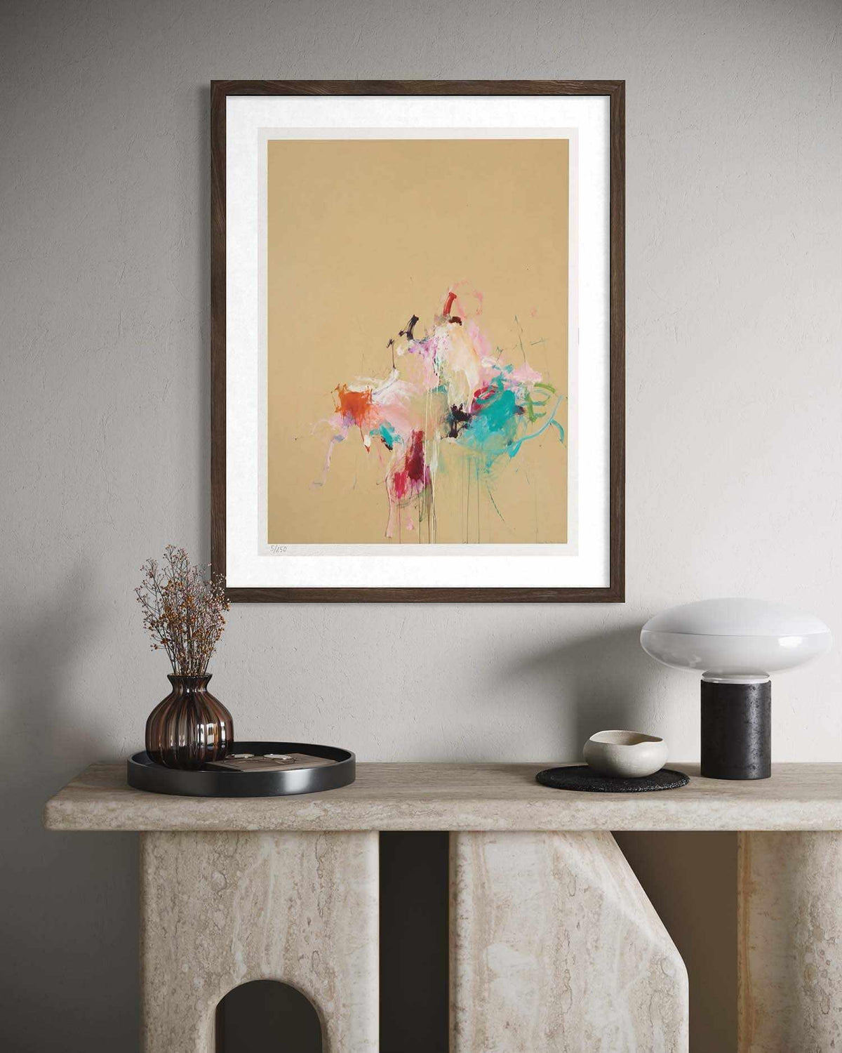 A framed abstract art print featuring colorful paint splatters on a warm beige background, styled on a stone console table against a gray wall.