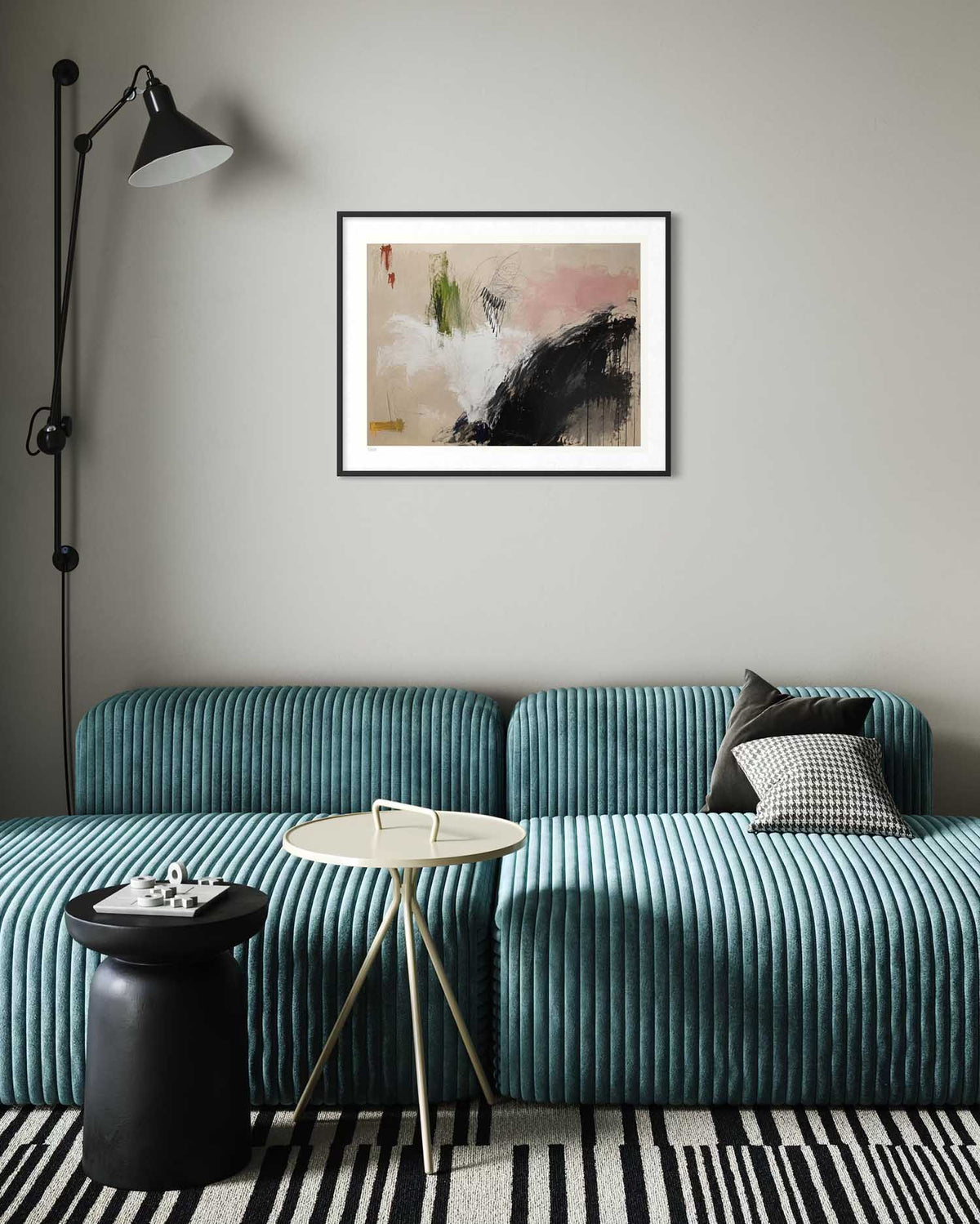 A framed abstract art print with black, white, pink, and green brushstrokes hangs on a light gray wall above a teal ribbed sofa in a modern living room setting.