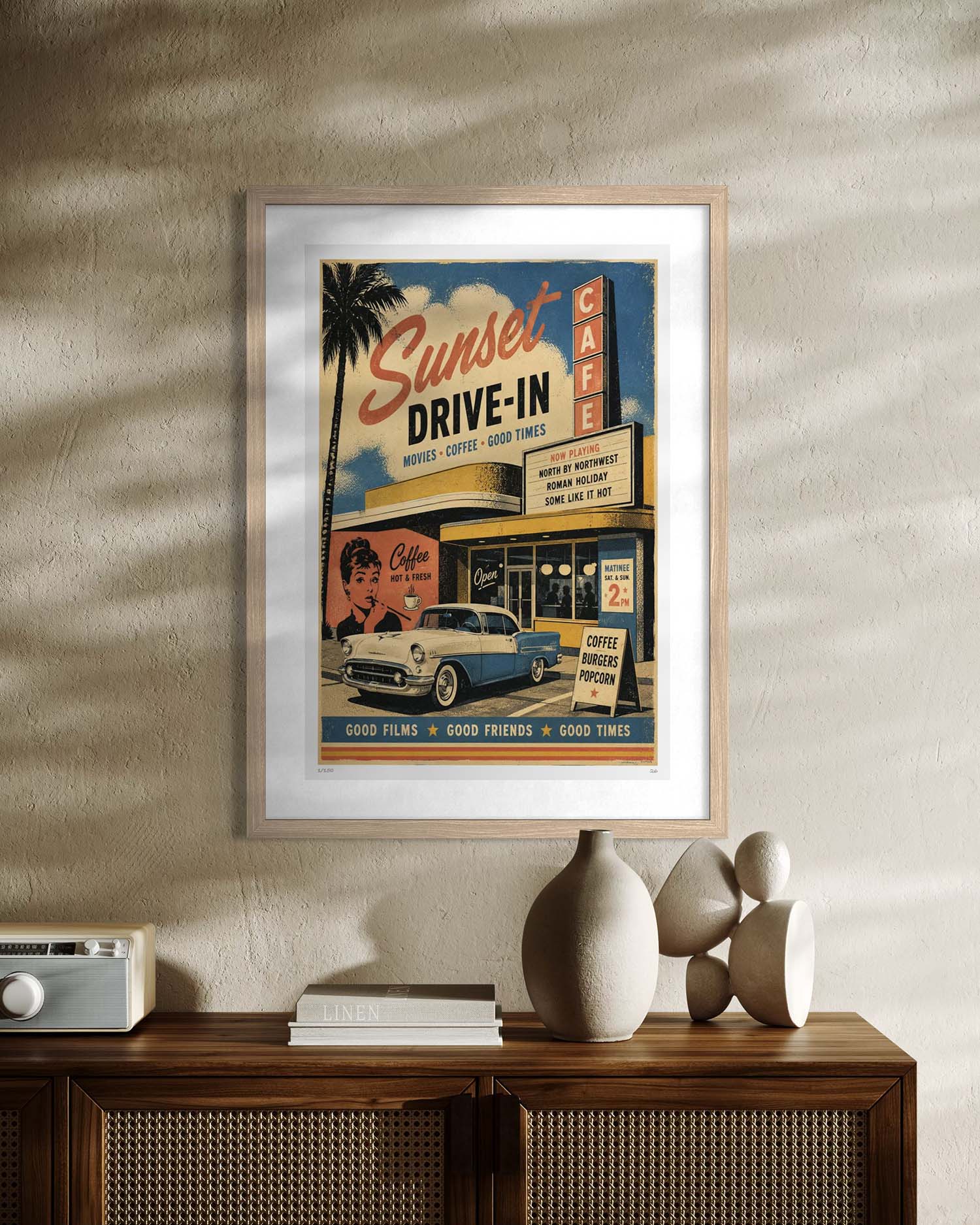 A framed vintage-style movie poster reading 'Sunset Drive-In' hangs on a textured beige wall above a wooden cabinet with decorative objects. The poster features retro cafe signage, a palm tree, faces, and a blue-and-white car, with a light wood frame and white mat.