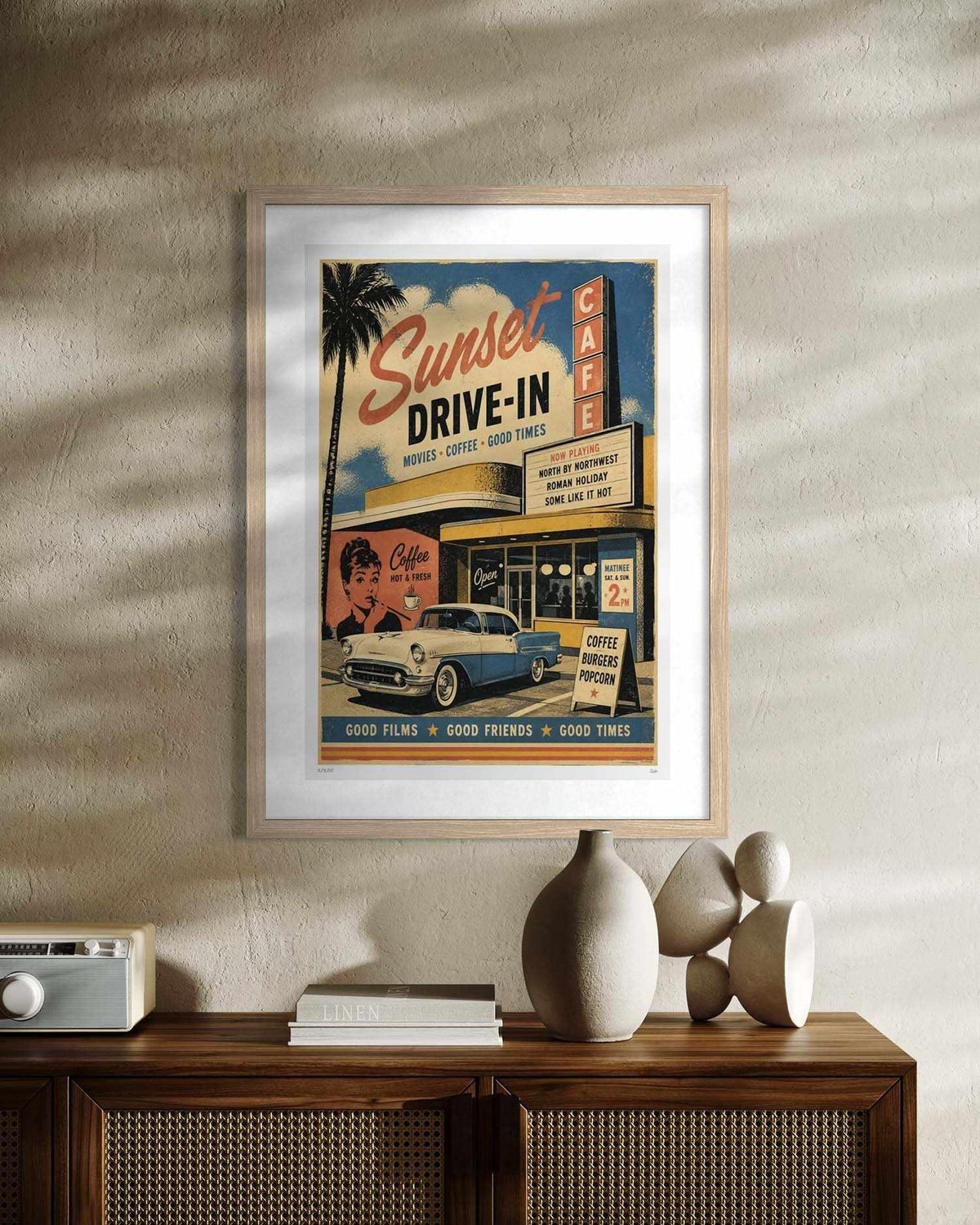 A framed vintage-style movie poster reading 'Sunset Drive-In' hangs on a textured beige wall above a wooden cabinet with decorative objects. The poster features retro cafe signage, a palm tree, faces, and a blue-and-white car, with a light wood frame and white mat.