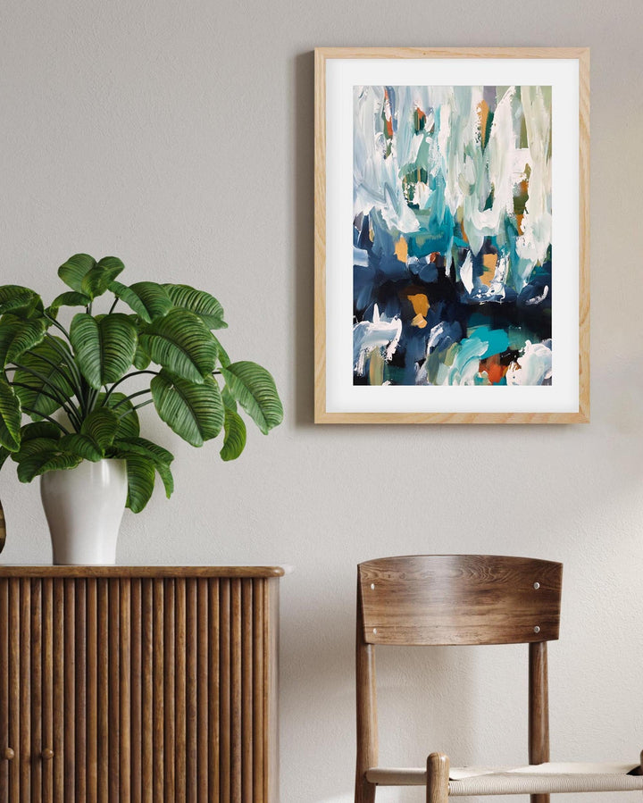 Framed abstract art on a wall above a wooden cabinet with a plant and chair.