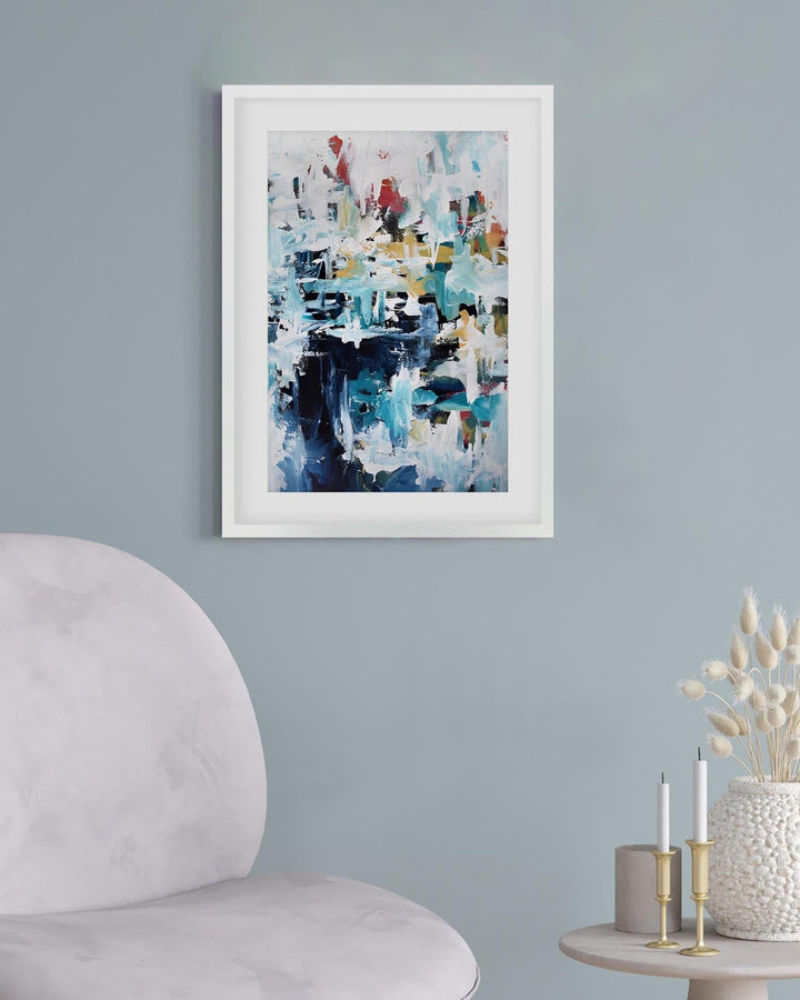 Framed abstract art piece on a light blue wall with a chair and small table in the foreground.