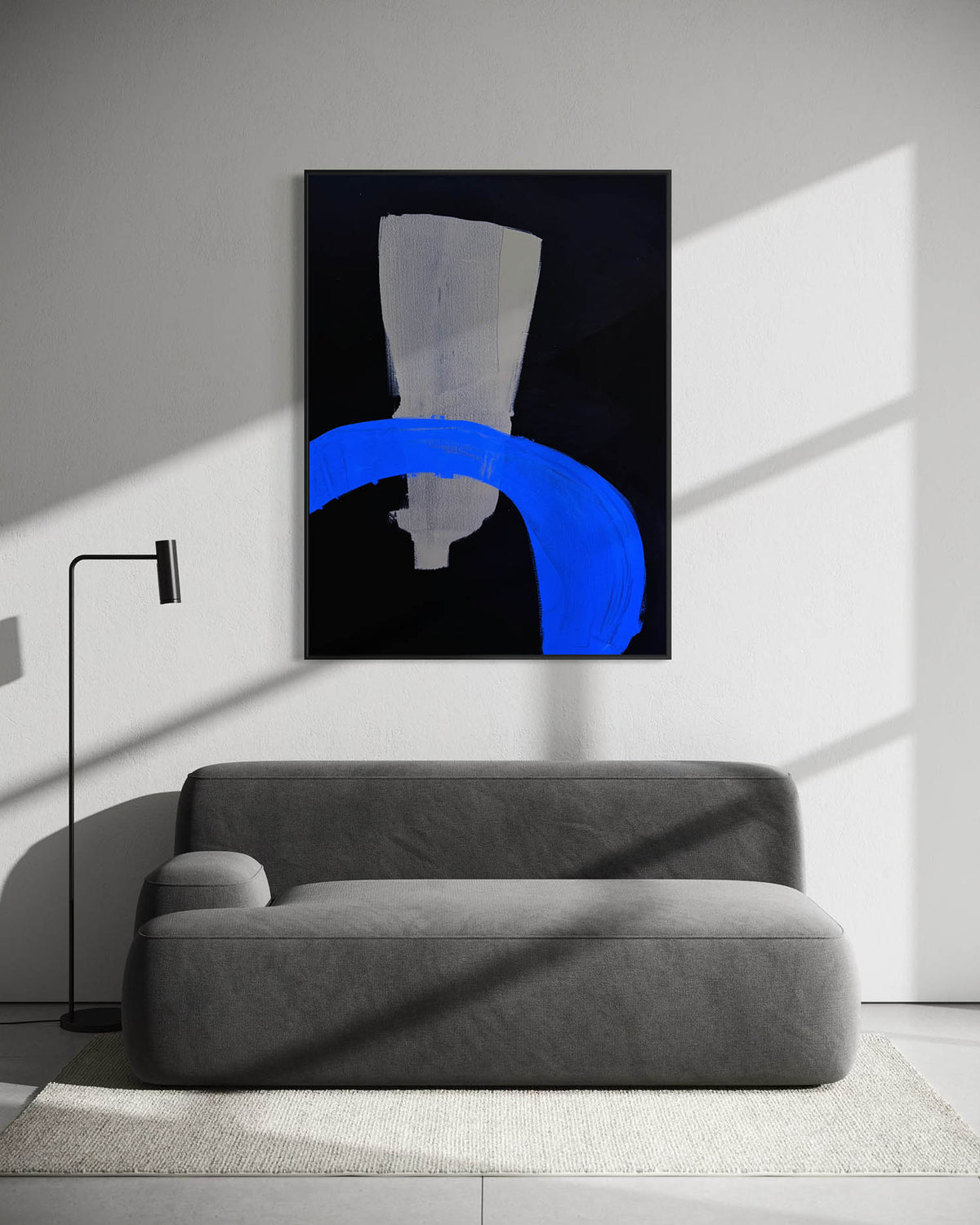 A framed abstract wall art print hangs above a gray sofa in a modern room with strong diagonal sunlight and shadows. The artwork features a black background with a large blue curved shape and a white geometric form.