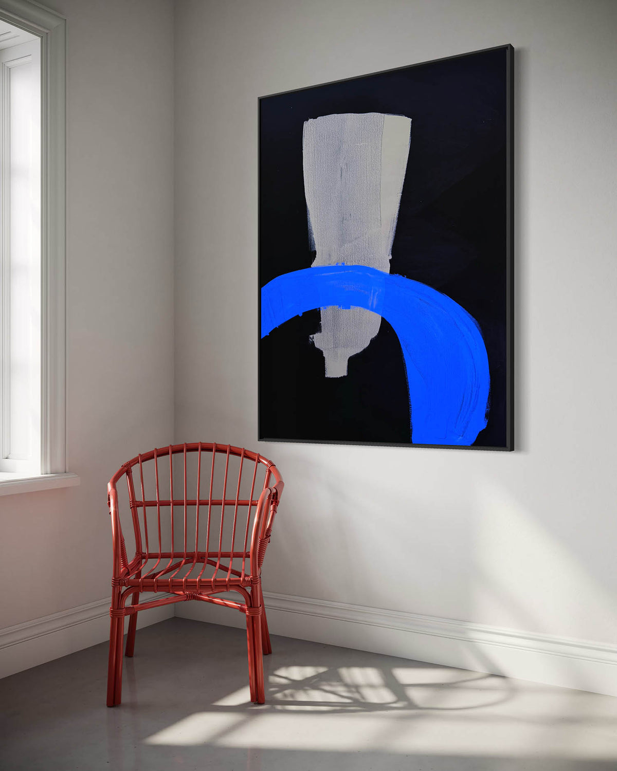 A large framed abstract canvas print hanging on a white wall in a bright room, featuring a black background with a white textured shape and a bold blue brushstroke. A red wire chair sits below the artwork near a window.