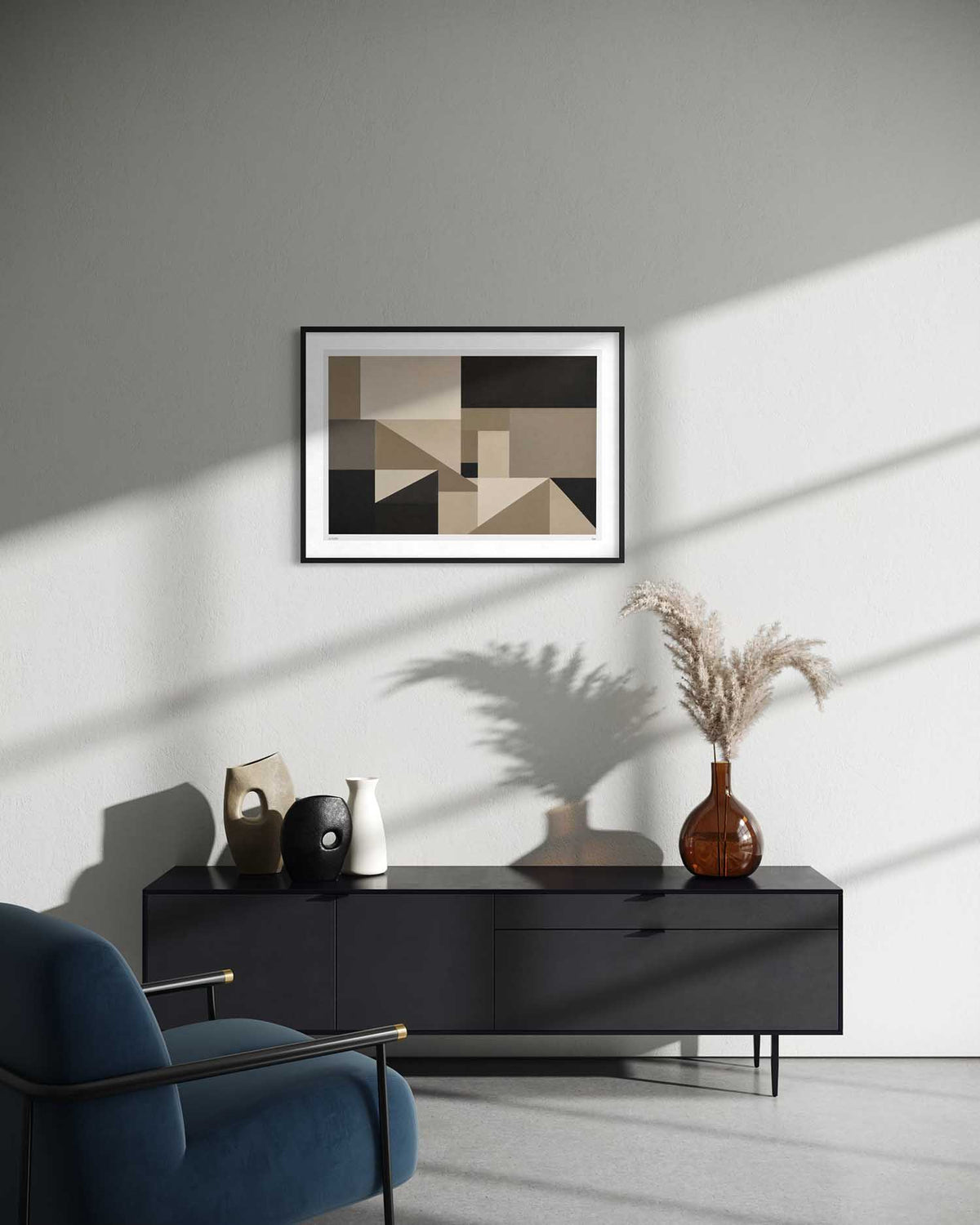 A framed abstract geometric art print in black, beige, and brown hangs on a light gray wall above a dark sideboard in a modern living room setting with a blue armchair and decorative vases.