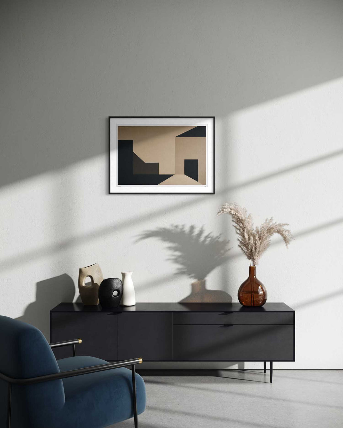 A framed abstract geometric art print with beige, black, and brown shapes hangs on a light gray wall above a dark sideboard in a modern living room setting with a blue armchair and dried pampas grass.