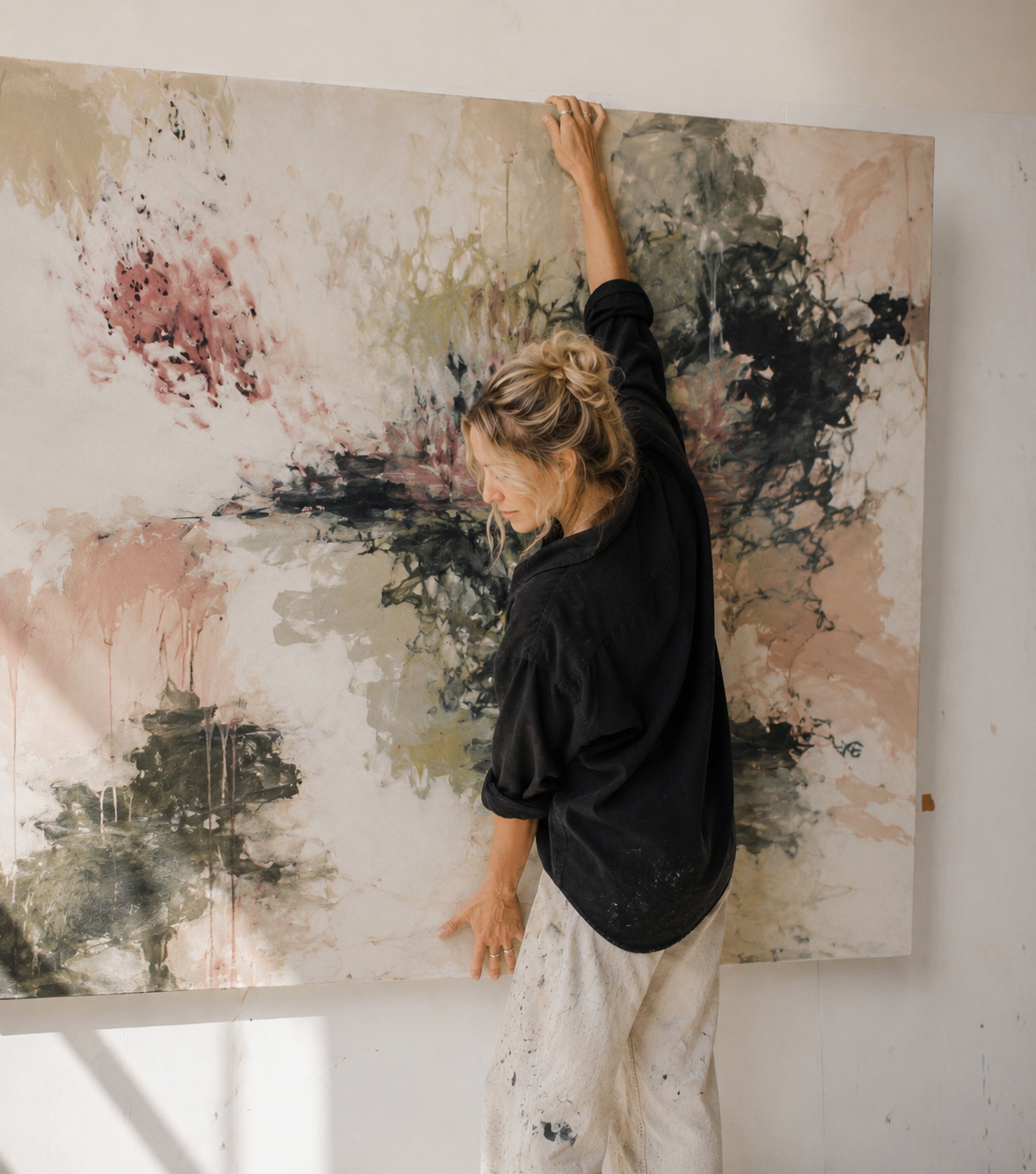 A woman in a black shirt and white pants stands beside a large abstract canvas painting with pink, black, and beige tones, holding the top edge of the artwork in a studio setting.