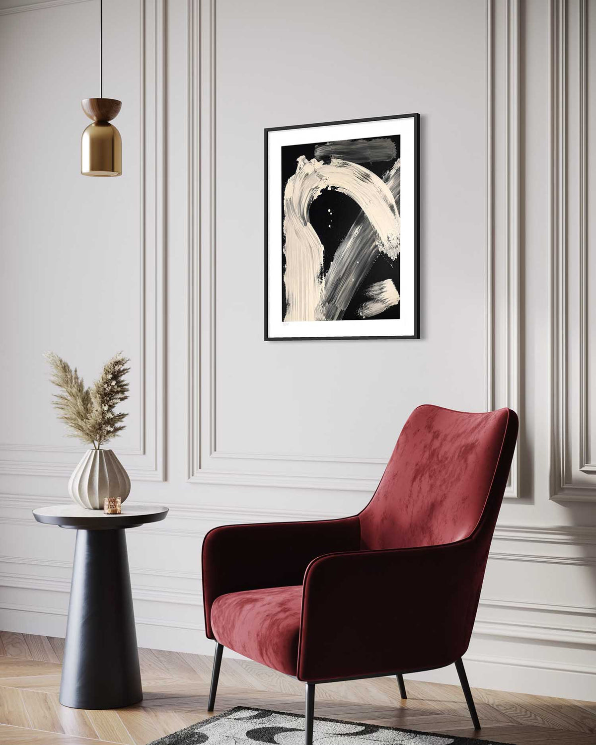 A framed abstract black and white art print hangs on a light gray paneled wall in a styled living room setting with a deep red velvet armchair, a small black side table with a vase of dried grass, and a hanging brass pendant light.