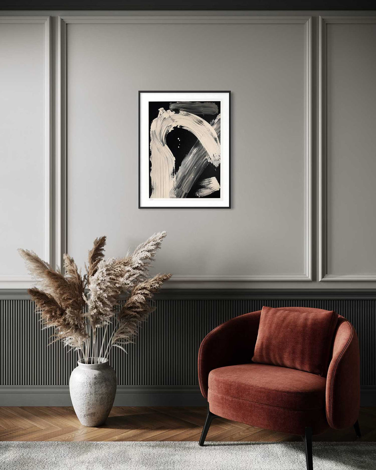 A framed abstract black and white art print hangs on a gray paneled wall above a rust-red armchair and a vase of dried pampas grass in a styled interior room.