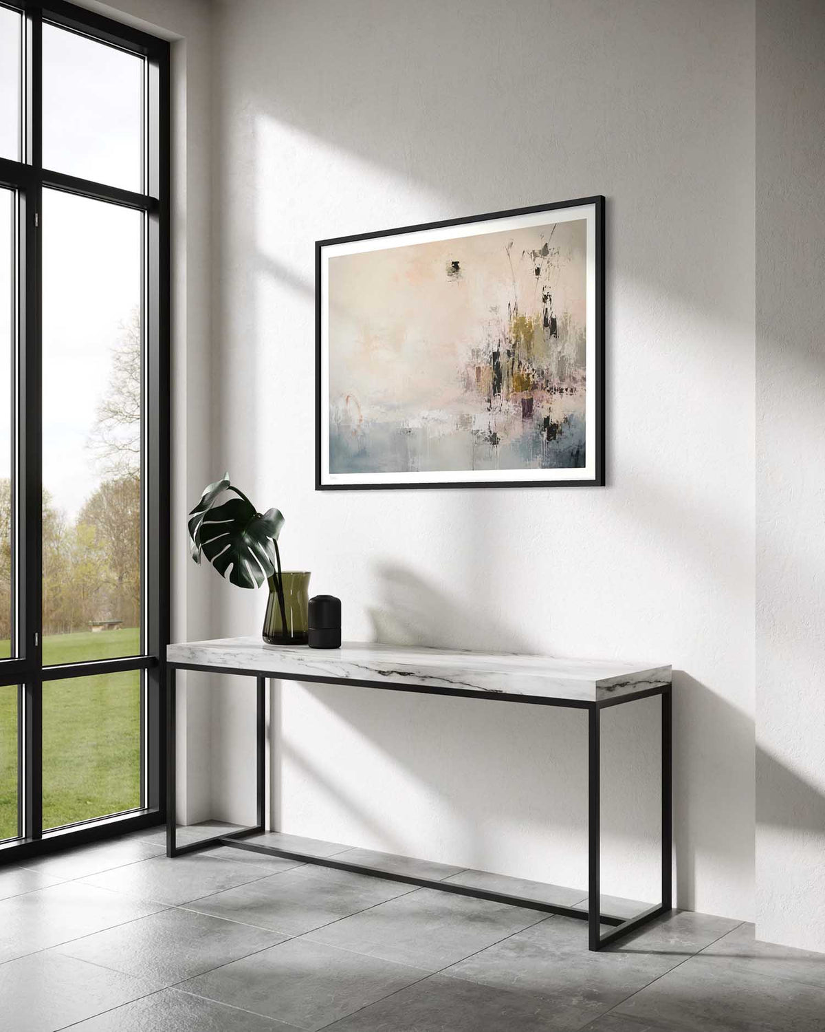 A framed abstract art print with a black frame hangs on a white wall above a modern console table in a bright, contemporary room with a large window.