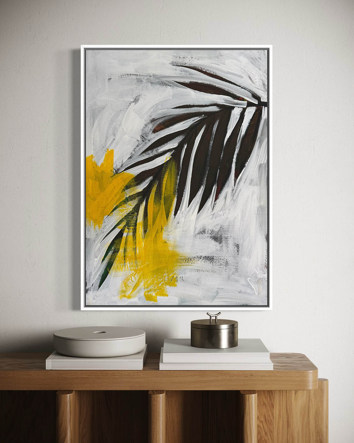 A framed abstract botanical wall art print featuring black and yellow brushstrokes on a white and gray background, hanging on a light gray wall above a wooden console table with books and decorative objects.