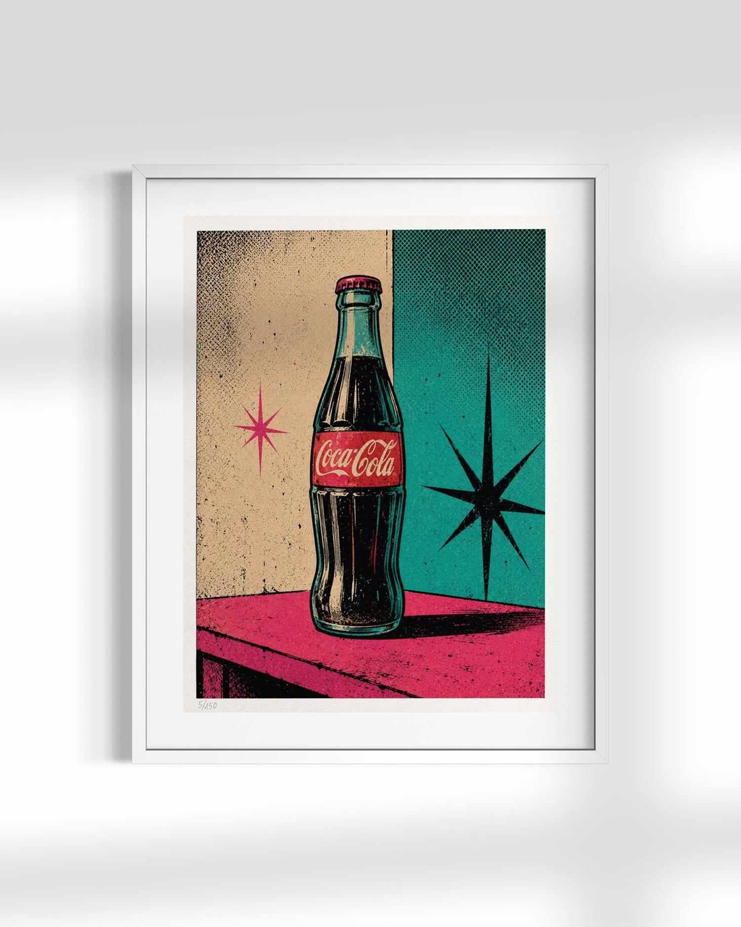 A framed pop-art print featuring a vintage Coca-Cola glass bottle on a pink table against a split beige and teal background with starburst shapes, shown against a light gray wall.