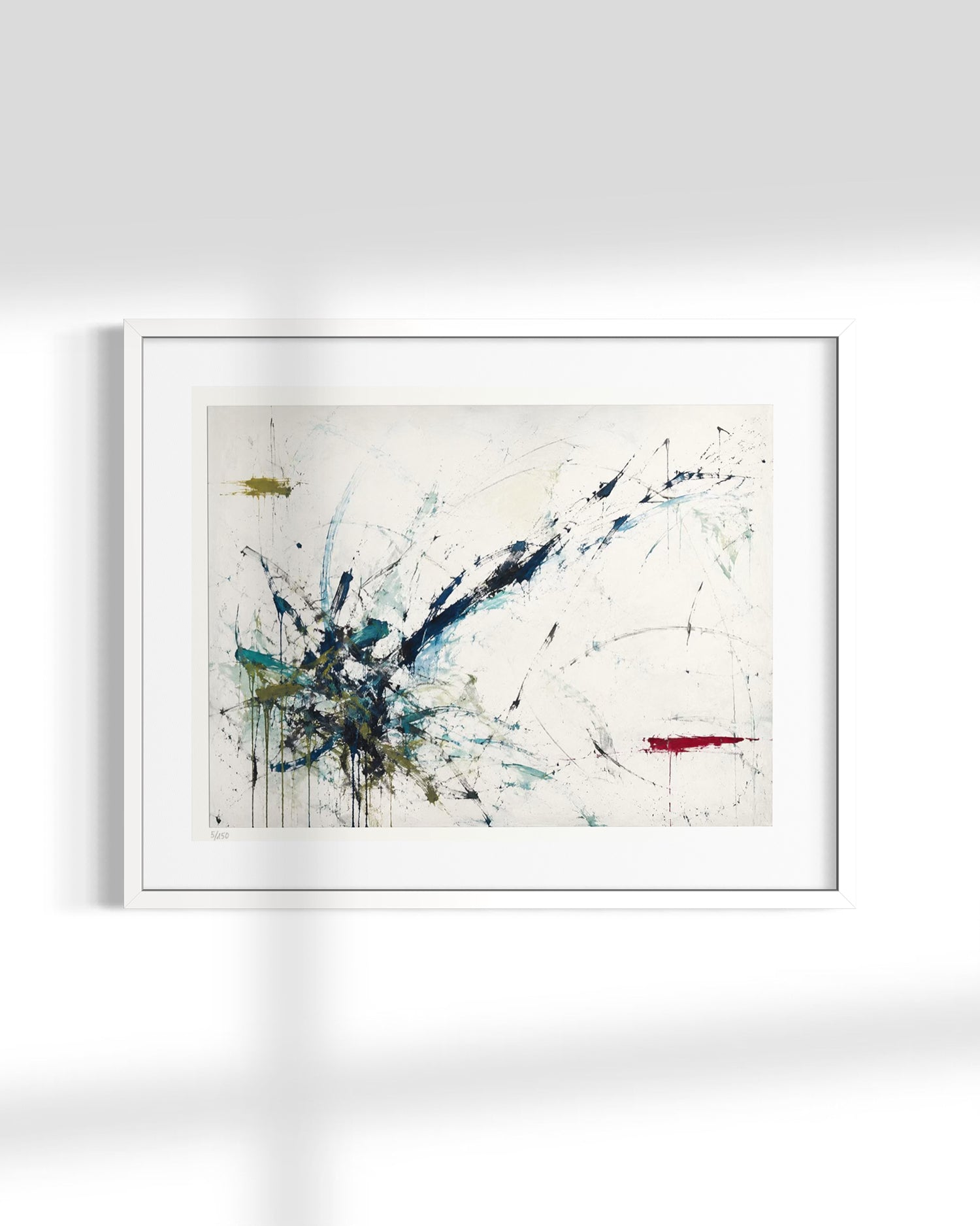 A framed abstract art print featuring expressive splatters and drips in blue, black, teal, and a small red stroke on an off-white background, displayed against a light gray wall with soft window-light shadows.
