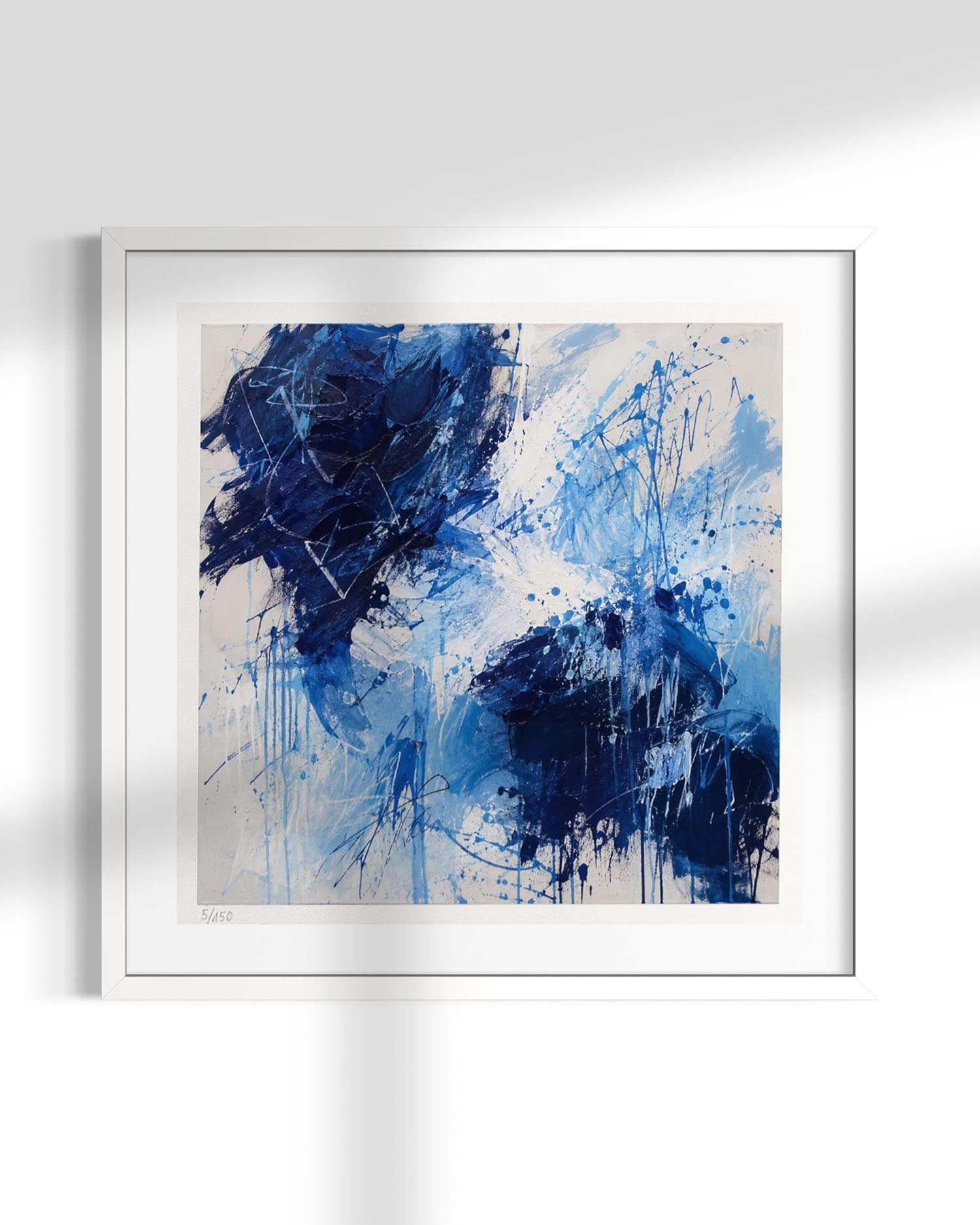 Abstract framed wall art featuring expressive blue and navy brushstrokes and paint splatters on a light beige background, presented in a white frame against a white wall with soft shadow overlays.