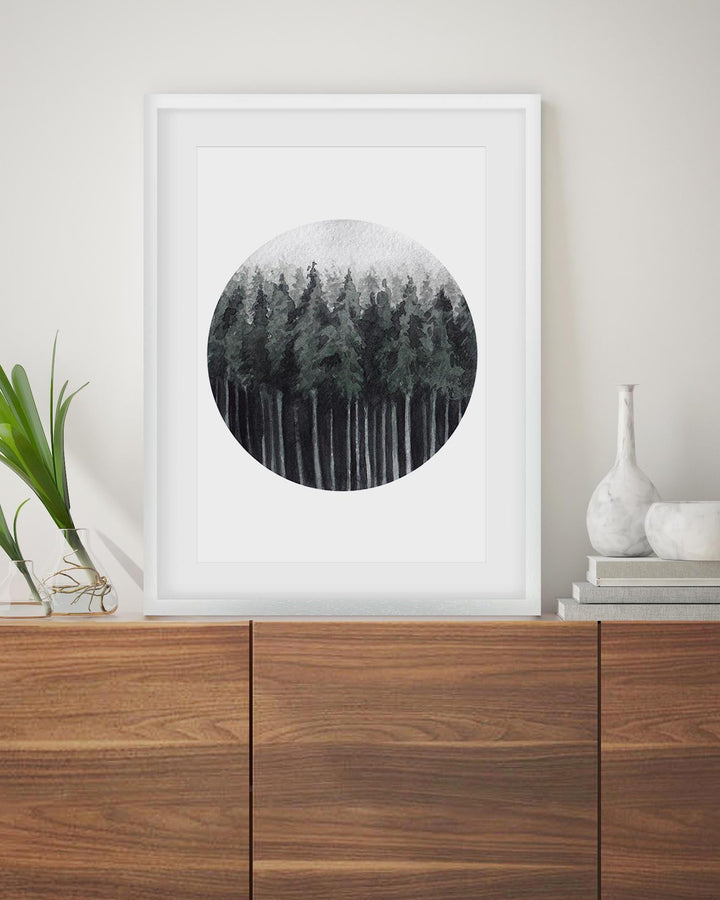 Framed circular artwork of a forest on a wall above a wooden cabinet.