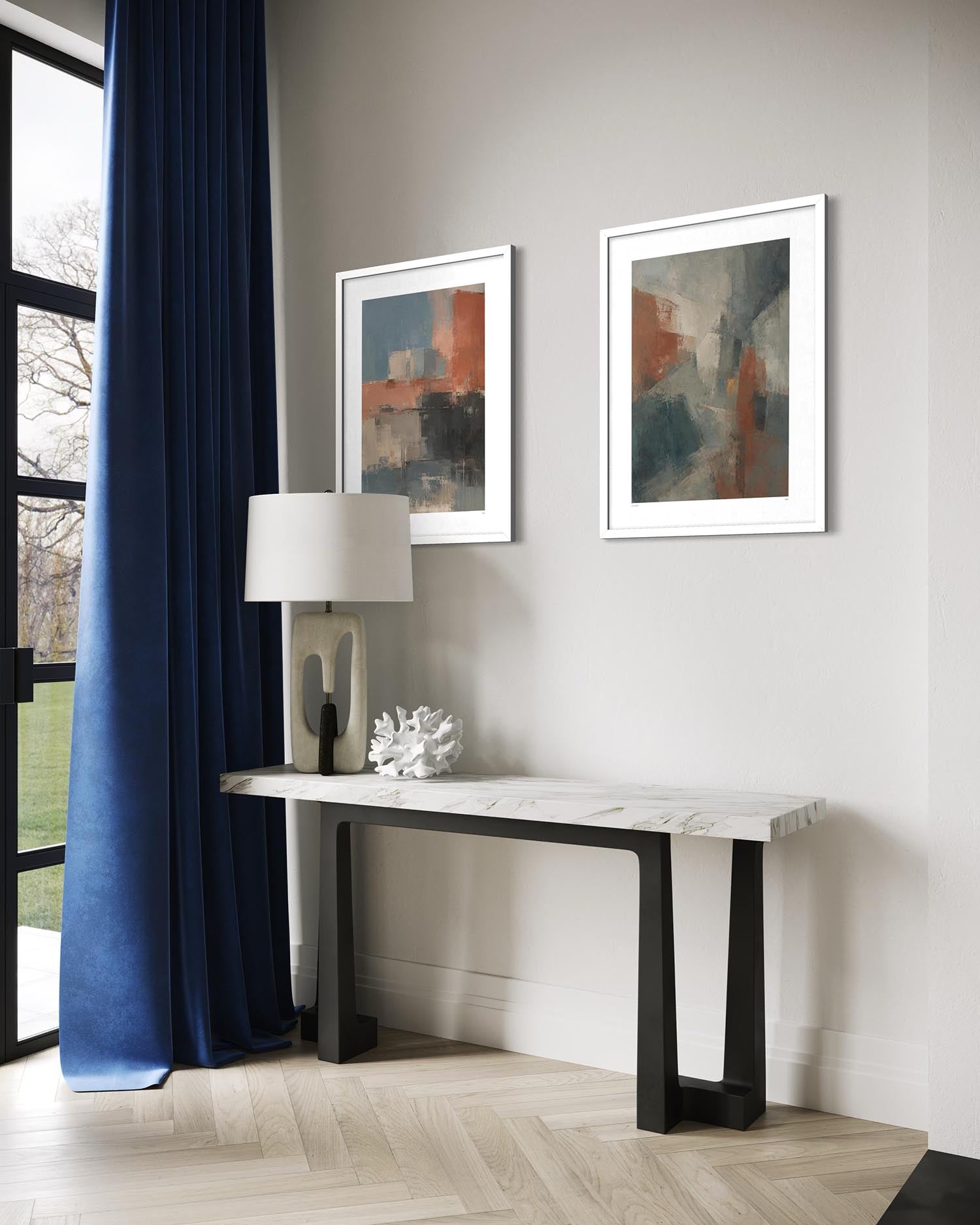 A styled interior scene showing two framed abstract paintings hanging on a light gray wall above a console table. The room includes a dark blue curtain, a table lamp, a small decorative object, wood flooring, and part of a window.