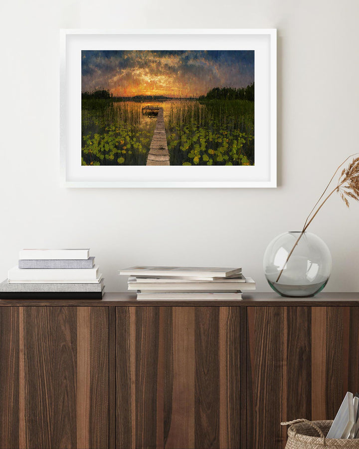 Framed artwork of a sunset over water on a wall above a wooden cabinet.