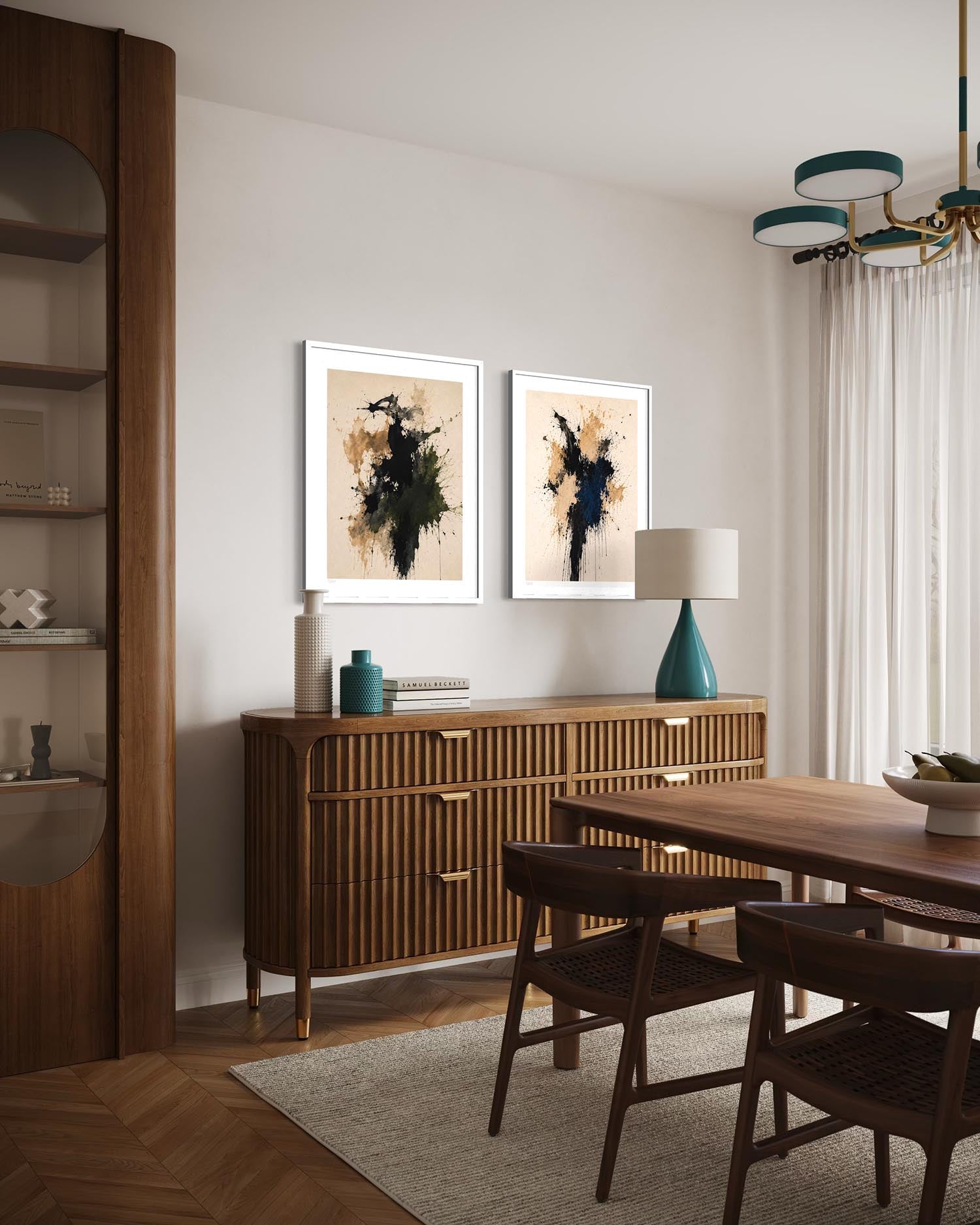 A styled dining room interior featuring two framed abstract art prints with black, white, and beige brushstrokes hanging above a mid-century modern wooden credenza. The room includes a wooden dining table and chairs, a teal ceramic lamp, and a tall wooden shelving unit on the left.