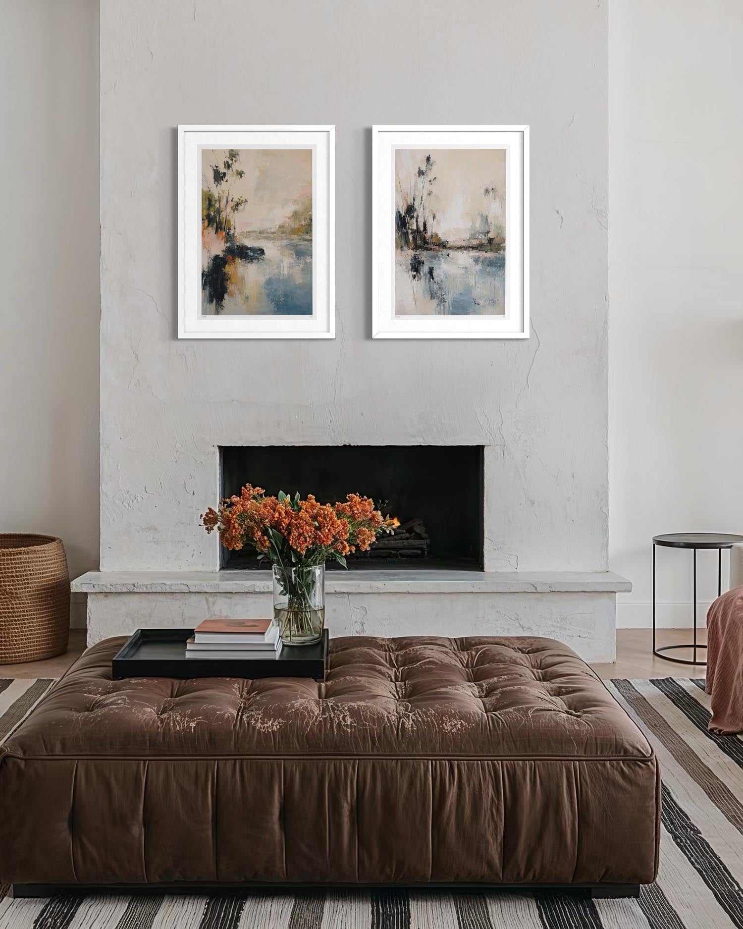 Two framed abstract landscape prints with blue, beige, and gray tones are displayed side-by-side above a fireplace in a modern living room setting with a brown tufted ottoman and orange flowers in the foreground.