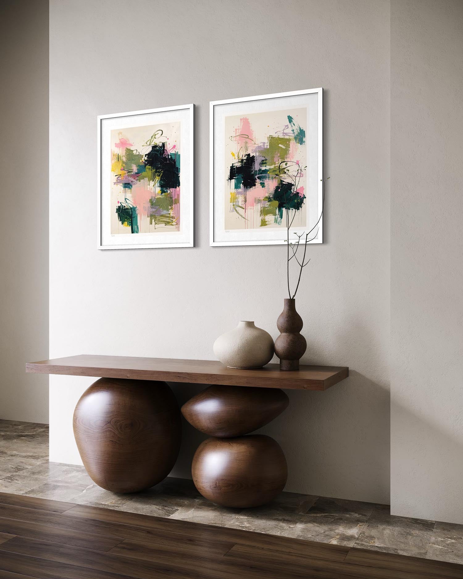 Two framed abstract art prints with pink, green, and black brushstrokes hang on a beige wall above a wooden console table with sculptural round bases and decorative vases.